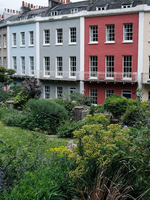 A city garden located in front of a multi-storey residential building painted in pastel blue and red, featuring a variety of lush green plants, shrubs, and flowering perennials arranged in bed borders