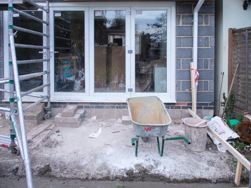 A garden patio area undergoing construction or renovation, featuring a partially finished concrete surface with scattered debris such as paper and small stones. A wheelbarrow filled with dirt, a woode